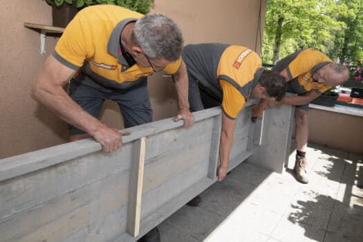 Werken in de ouderenzorg is nooit makkelijk geweest, maar de pandemie was een ongekende uitdaging voor het personeel. Op 12 mei, de wereldwijde Dag van de Verpleging, kregen de medewerkers van Attent Zorg en Behandeling in Dieren daar alle lof voor. De directie wilde haar waardering graag vertalen in een concreet gebaar, maar helaas ontbrak het daarvoor aan budget. HORNBACHhelpt kwam in actie: een verlept terras werd opgeknapt tot sfeervolle personeelsruimte, waar de medewerkers in alle rust kunnen opladen. Ook de bewoners, die elkaar én het personeel door de zware tijd hebben gesleept, kregen een prachtige nieuwe plek. Attent Zorg en Behandeling biedt ouderen zorg op maat, aan huis of op een van de elf locaties in Gelderland. Op de woonzorglocatie in Dieren, De Oude Plataan, stonden op de Dag van de Verpleging de zorgprofessionals centraal. Dankzij de steun van HORNBACHhelpt kreeg hún dagelijkse werk een krachtige impuls, en ook de levens van de bewoners, die het de afgelopen tijd zwaar voor de kiezen hebben gekregen, werden letterlijk en figuurlijk opgefleurd. Langetermijnsteun "Zo'n Dag van de Verpleging is hartstikke mooi," zegt senior verpleegkundige Emmy Kreuzen, "maar ik vind het nóg belangrijker dat ons personeel die steun ook op de lange termijn voelt. Daar had onze directie met liefde zelf voor gezorgd, maar ons budget is volledig bestemd voor de ouderenzorg. Onze inkomsten komen vanuit de bewoners en zijn voor de bewoners. Dat betekent helaas dat er weinig overblijft voor de medewerkers. Dankzij HORNBACHhelpt hebben we toch iets belangrijks voor hen kunnen doen, vandaag en op de langere termijn. Dat is indirect al goed voor onze bewoners, omdat het personeel er dan nóg beter voor hen kan zijn, maar we vinden het helemaal geweldig dat HORNBACHhelpt de ouderen zelf óók gericht wilde helpen." Goede voorbereiding Vanaf tien uur 's ochtends nam een team van zeven HORNBACHhelpt-vrijwilligers meerdere plekken op De Oude Plataan onder handen. "Zowel voor de zorgmedewerkers als voor de bewoners hebben we een compleet nieuw terras gemaakt: twee rustige, comfortabele buitenruimtes, op plekken die er al tijden wat verlept bijlagen," zegt Peter Vuijk, vrijwilliger van HORNBACHhelpt. "De afgelopen weken hebben we alles opgemeten. Aan de hand daarvan zijn we vol enthousiasme grote banken, tafels en bloembakken gaan ontwerpen. Door al het voorwerk was het vandaag vooral een kwestie van in elkaar zetten en bevestigen. Dat ging soepel, in een bijna feestelijke sfeer. Je merkte echt dat de bewoners en het personeel in het zonnetje werden gezet - en terecht!" Kwaliteit van leven Naast de nieuwe personeelslounge buiten, werd ook het balkon op de eerste verdieping aangepakt. Ook daar werden fleurige bloembakken en planten geplaatst, maar ook zitbanken en verrijdbare tafels. Door de aangepaste hoogte kunnen ook de bewoners met rolstoelen er makkelijk aanschuiven. "Ze hebben nu een extra mogelijkheid om heel gemakkelijk van de buitenlucht te genieten," zegt Emmy Kreuzen. "Het is een fijne, uitnodigende plek geworden, waar onze bewoners lekker met een klein groepje in de schaduw kunnen zitten. Dat is zeker in deze warme tijd van het jaar een flinke impuls voor hun gezondheid, én voor hun kwaliteit van leven." Frisse moed Met een paar uur stevig klussen steunde HORNBACHhelpt in één klap tientallen bewoners en medewerkers, en dat is zeer welkom, benadrukt Emmy. "In de ouderenzorg moet je vaak ontzettend schakelen. Het ene moment breng je een kopje koffie, en dan zit je opeens bij een sterfgeval. Dat vergt natuurlijk veel van onze medewerkers én van de ouderen zelf. De afgelopen periode was voor ons allemaal best zwaar. Dan zijn zulke plekken om dagelijks even tot rust te komen niet alleen fijn: ze zijn ook echt nodig. Dankzij HORNBACHhelpt kunnen we er met z'n allen, ook na deze speciale dag, met frisse moed weer tegenaan!"